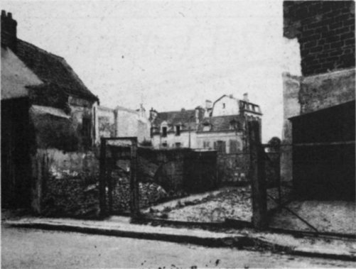 Vue d'une cour donnant sur la rue du Métier. Elle est grillagée et encadrée de maisons. On voit au fond les hauts bâtiments desservis par d'autres rues.
